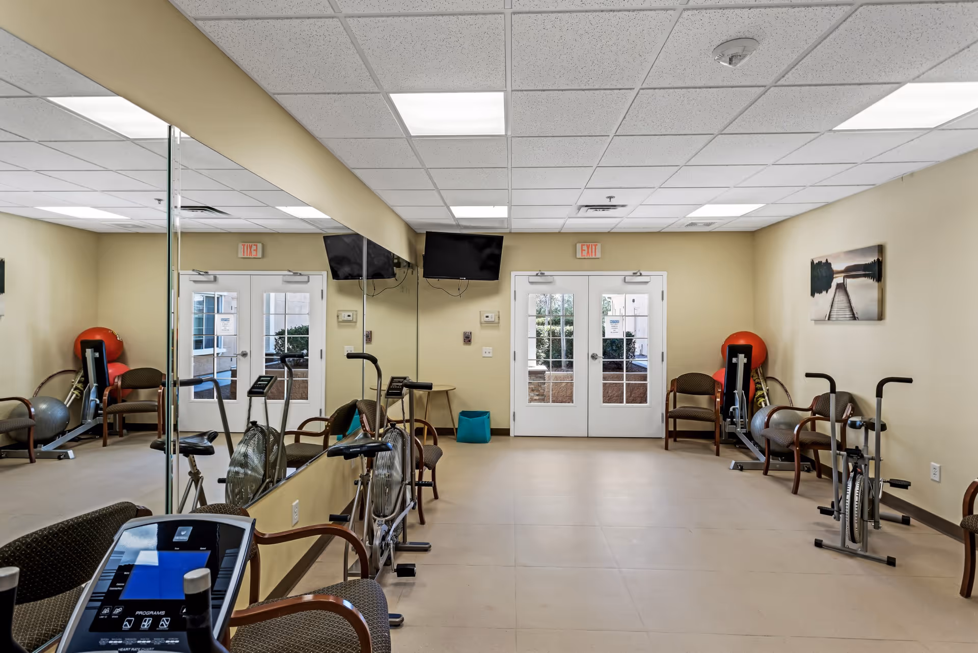 A small exercise room with stationary bikes, chairs, exercise balls, and a rowing machine. The room has beige walls, a tiled floor, a large mirror on one wall, and double glass doors leading outside. Two flat-screen TVs are mounted on the wall above the doors, and a framed picture of a dock over water hangs on the right wall.