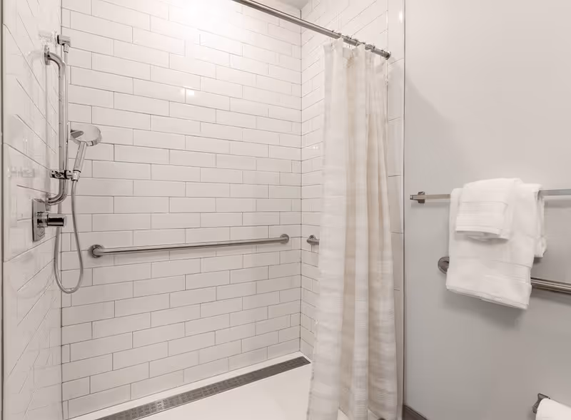 A white tiled accessible shower with grab bars, a handheld showerhead, and a shower curtain with towels on a wall-mounted rack.