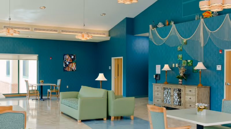 A bright and spacious common area with teal walls and light-colored flooring. The room features green cushioned sofas, wooden chairs, and tables. Decorative elements include two table lamps on a wooden sideboard, wall art, and hanging net decorations. Large windows allow natural light to enter the space.