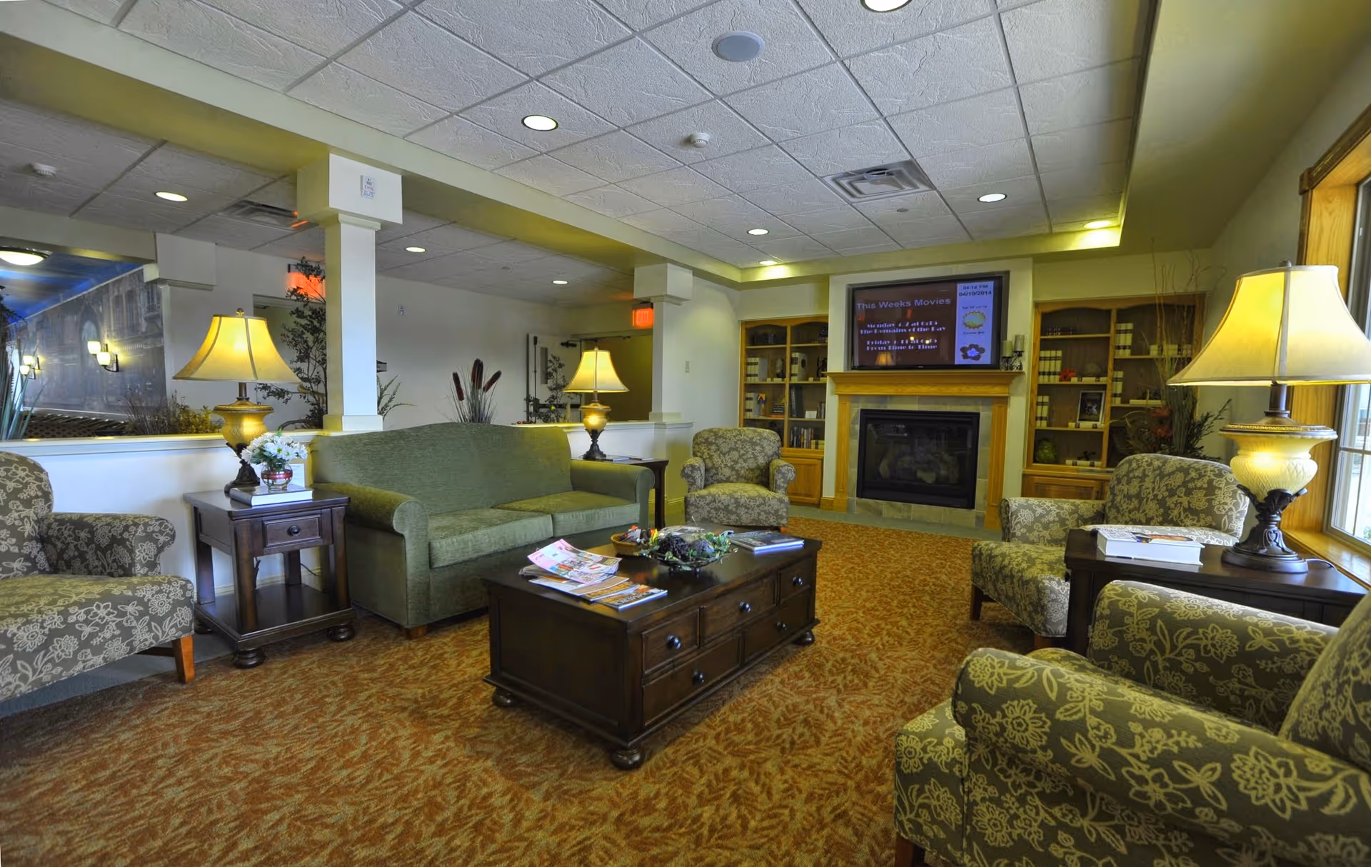 A cozy living room area in a retirement community featuring a green sofa, four patterned armchairs, two wooden side tables with lamps, a wooden coffee table with magazines, a fireplace with built-in shelves, and a TV mounted above the fireplace displaying movie information.