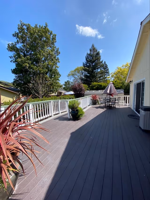 A spacious outdoor deck area with dark wooden flooring, white railing, and several potted plants. There is a patio table with chairs and a closed umbrella. The deck is attached to a light-colored building, and trees and a clear blue sky are visible in the background.