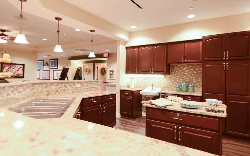A spacious kitchen area with dark wooden cabinets, granite countertops, a double sink, and various kitchen items such as bowls and containers. The kitchen is well-lit with pendant lights hanging from the ceiling and has a warm, inviting atmosphere. In the background, there is a hallway with framed pictures on the wall and a ceiling fan with lights.