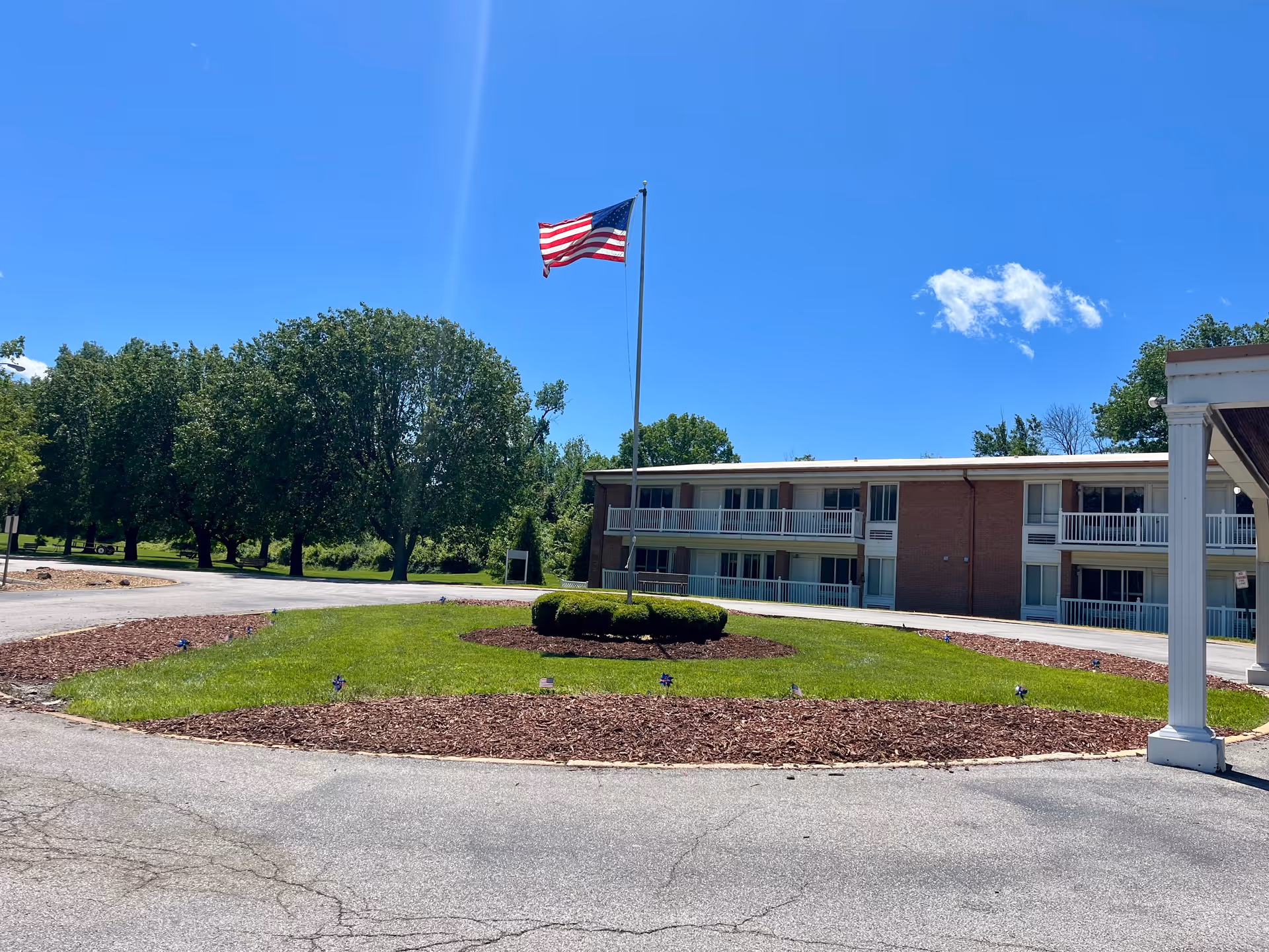 Exterior view of Athene Nursing and Rehabilitation facility on a sunny day with a clear blue sky. The image shows a two-story brick building with balconies, a circular driveway with a landscaped island in the center featuring trimmed bushes and an American flag on a flagpole. Trees and greenery are visible in the background.
