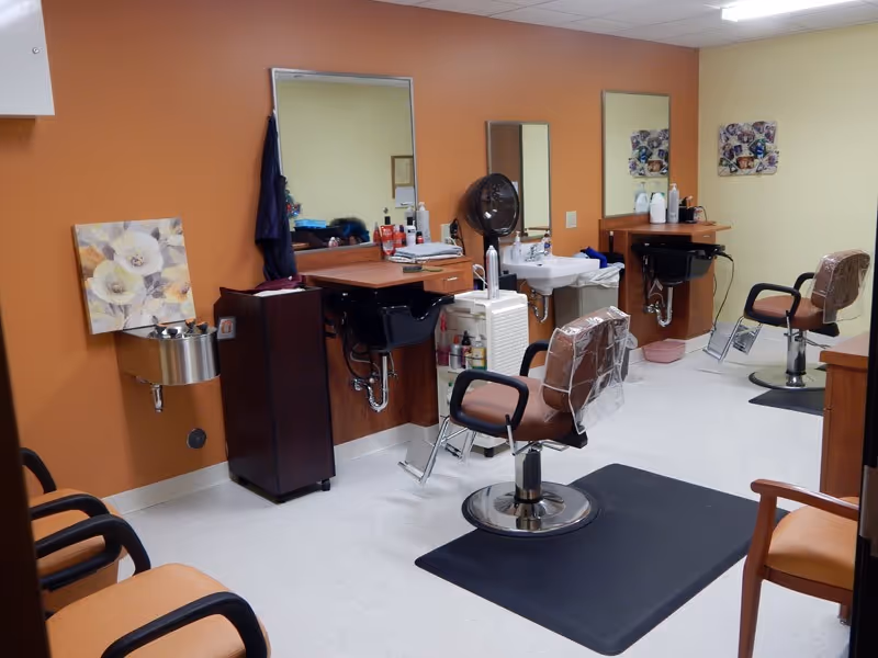 Interior view of a hair salon area in a care facility with two salon chairs in front of mirrors and sinks, various hair care products on counters, and chairs along the wall for waiting.