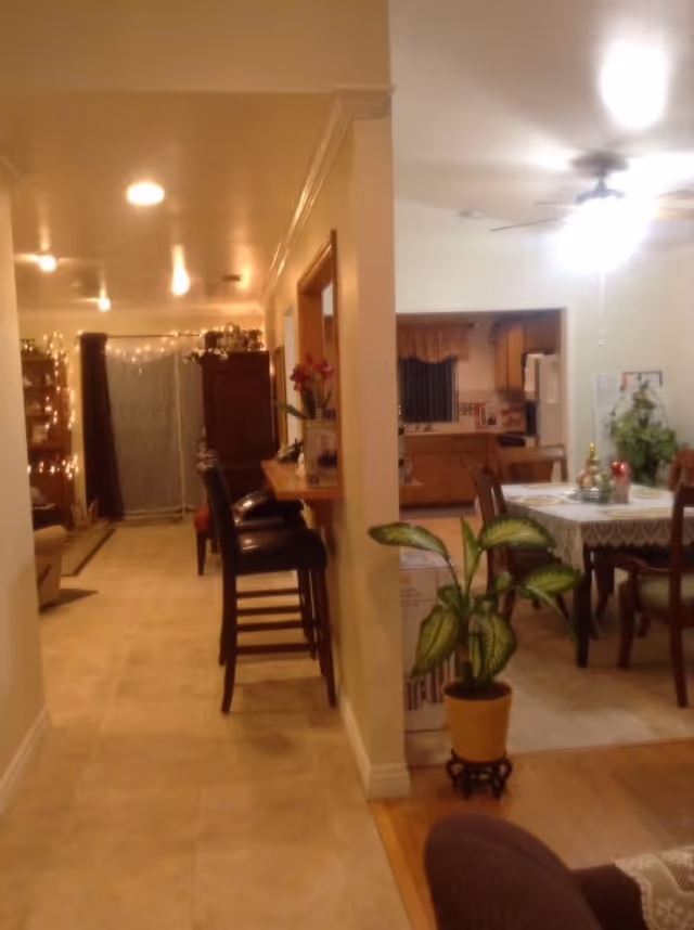 Interior view of a senior living facility showing a hallway with bar stools on the left and a dining area with a table and chairs on the right. The dining area has a ceiling fan with light, a potted plant near the entrance, and a kitchen visible in the background. The hallway is decorated with string lights and furniture.