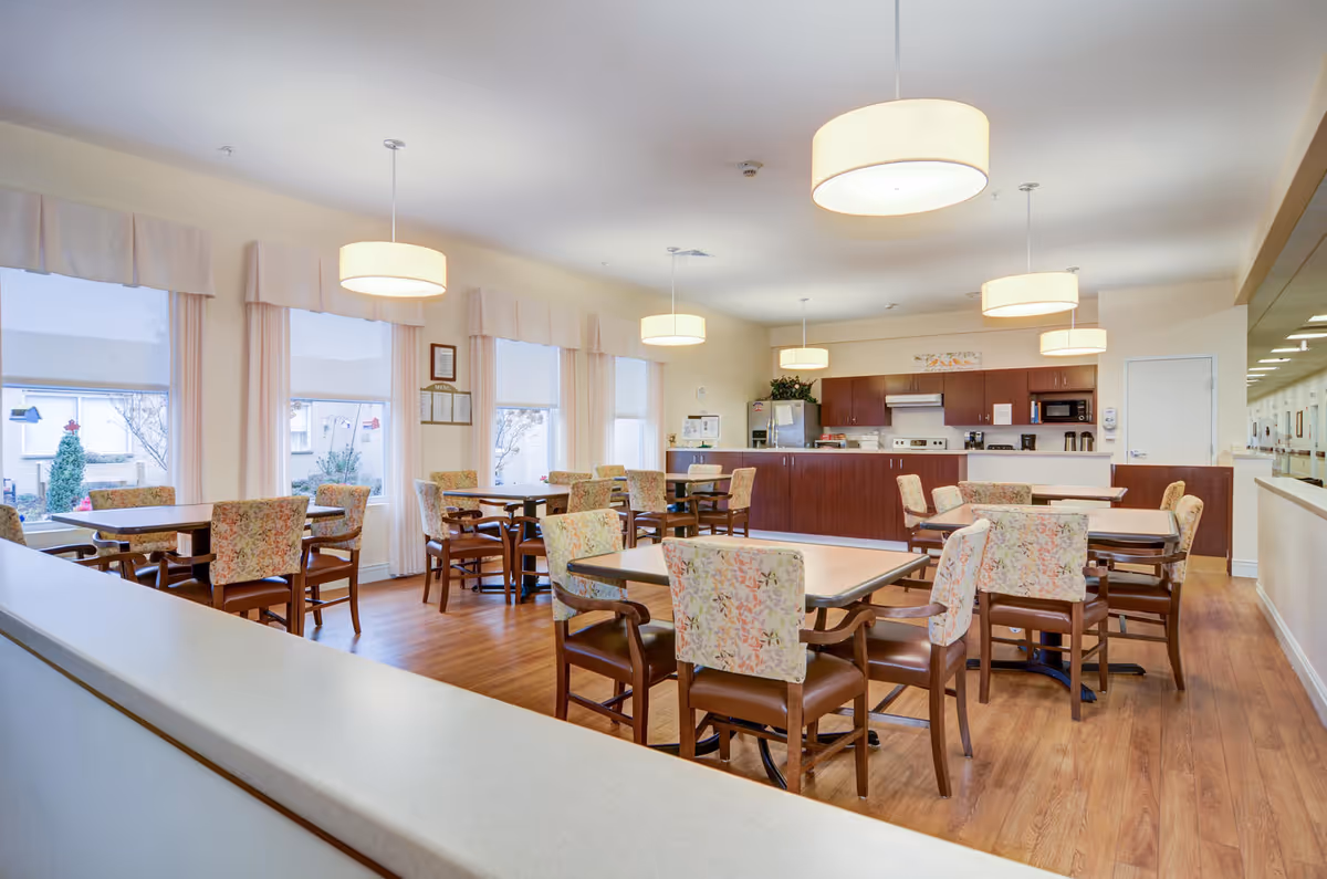 Bright dining room with multiple tables and floral-upholstered chairs, pendant lights, and a kitchen counter in the back.