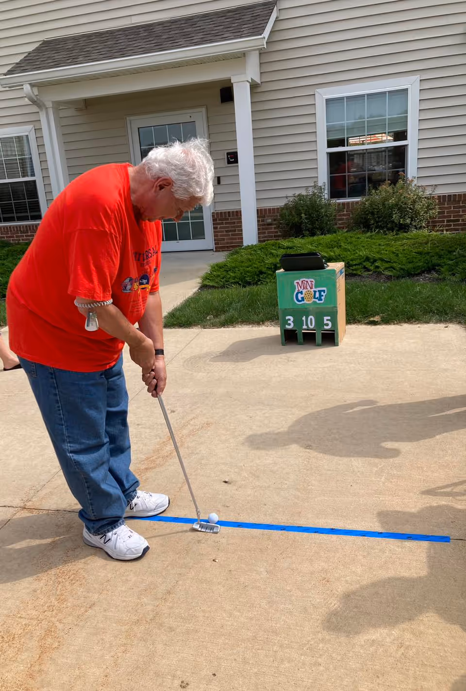 An elderly man wearing a red shirt and blue jeans is playing mini golf on a concrete path outside a building. He is holding a putter and aiming at a golf ball on the ground near a blue line. Behind him, there is a mini golf scoreboard and some green bushes in front of a beige building with white trim and windows.