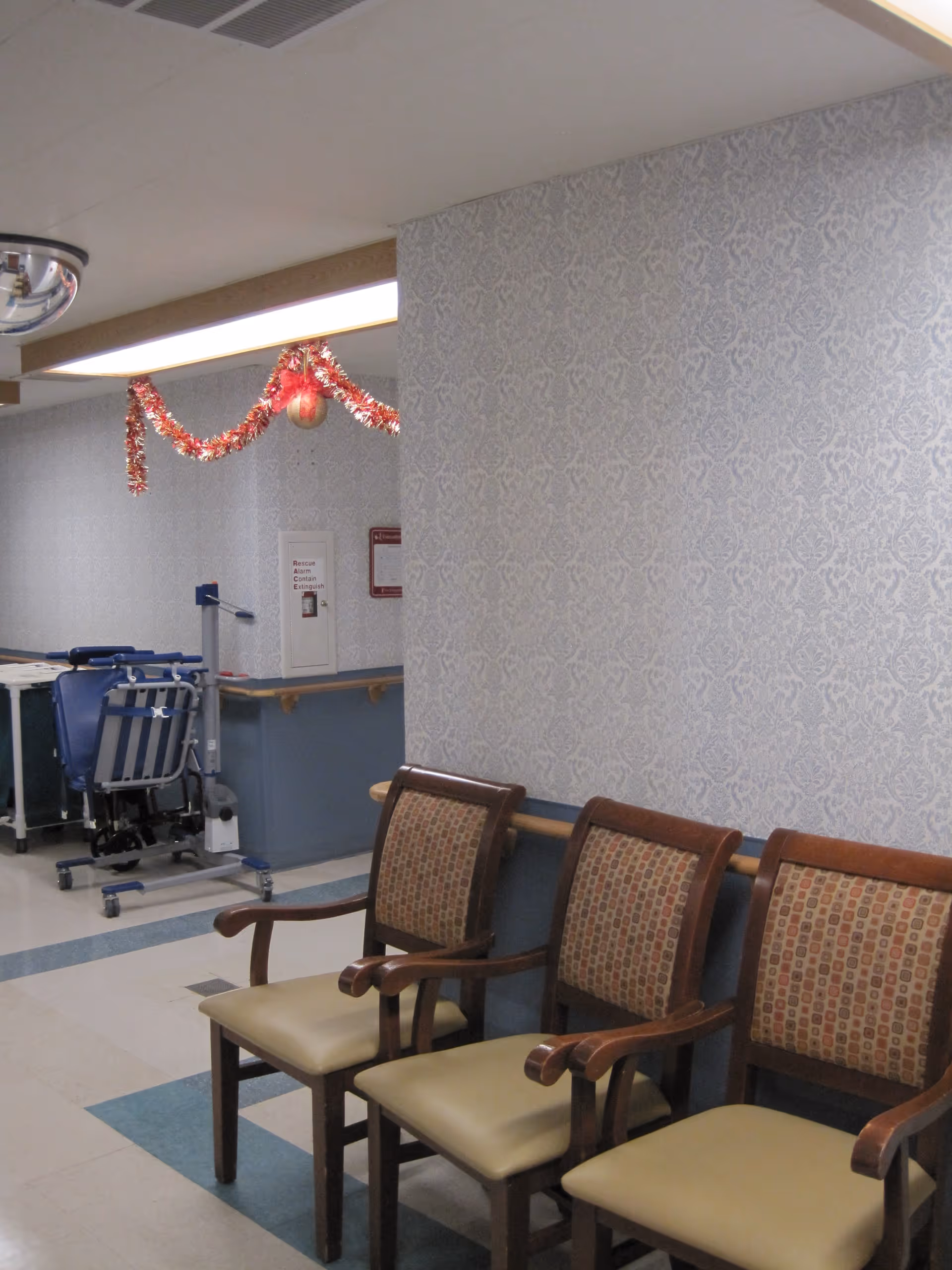 A hallway area in a senior care facility with three wooden chairs with patterned cushions lined up against a wall with light blue patterned wallpaper. There is a handrail along the wall and a ceiling light fixture. In the background, there is medical equipment and a fire extinguisher cabinet. Red and gold holiday garland is hanging from the ceiling.