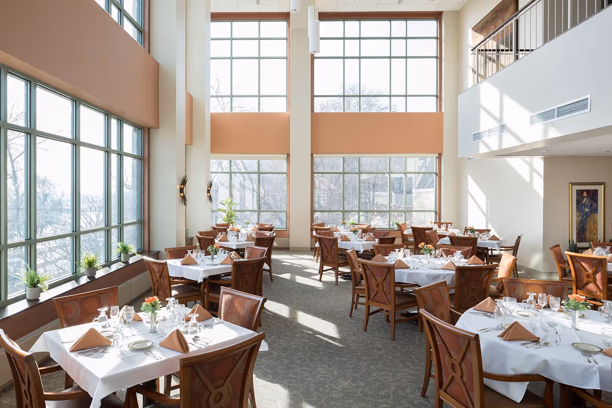 Bright double-height dining room with large windows and multiple tables set with white linens and place settings.