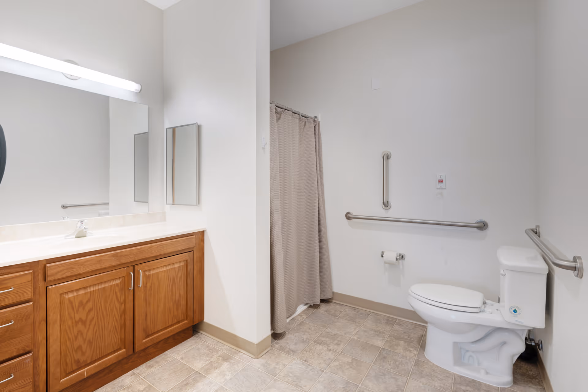 A clean and accessible bathroom with a wooden vanity cabinet, a large mirror, a sink, a toilet with grab bars on both sides, a toilet paper holder, and a shower area with a beige curtain.