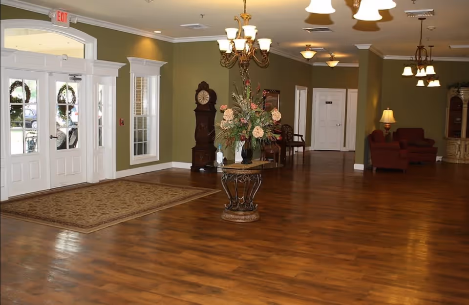 Spacious interior lobby area of Stonecrest Assisted Living Facility with wooden flooring, green walls, a decorative round table with a floral arrangement in the center, a grandfather clock against the wall, multiple ceiling light fixtures, a seating area with red chairs and a lamp, and large white double doors with glass panels leading outside.