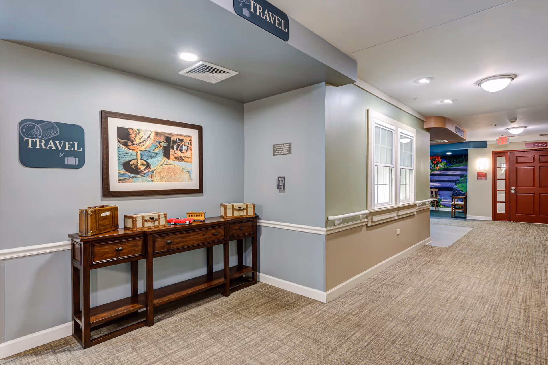 Interior hallway of a senior living facility with beige carpet and light-colored walls. On the left side, there is a wooden console table with decorative vintage suitcases and a small toy car on top. Above the table is a framed picture of a globe and travel-related items, along with a sign that says 'TRAVEL'. The hallway has handrails along the walls and leads to a red door at the end. There are windows on the right side and ceiling lights illuminating the space.
