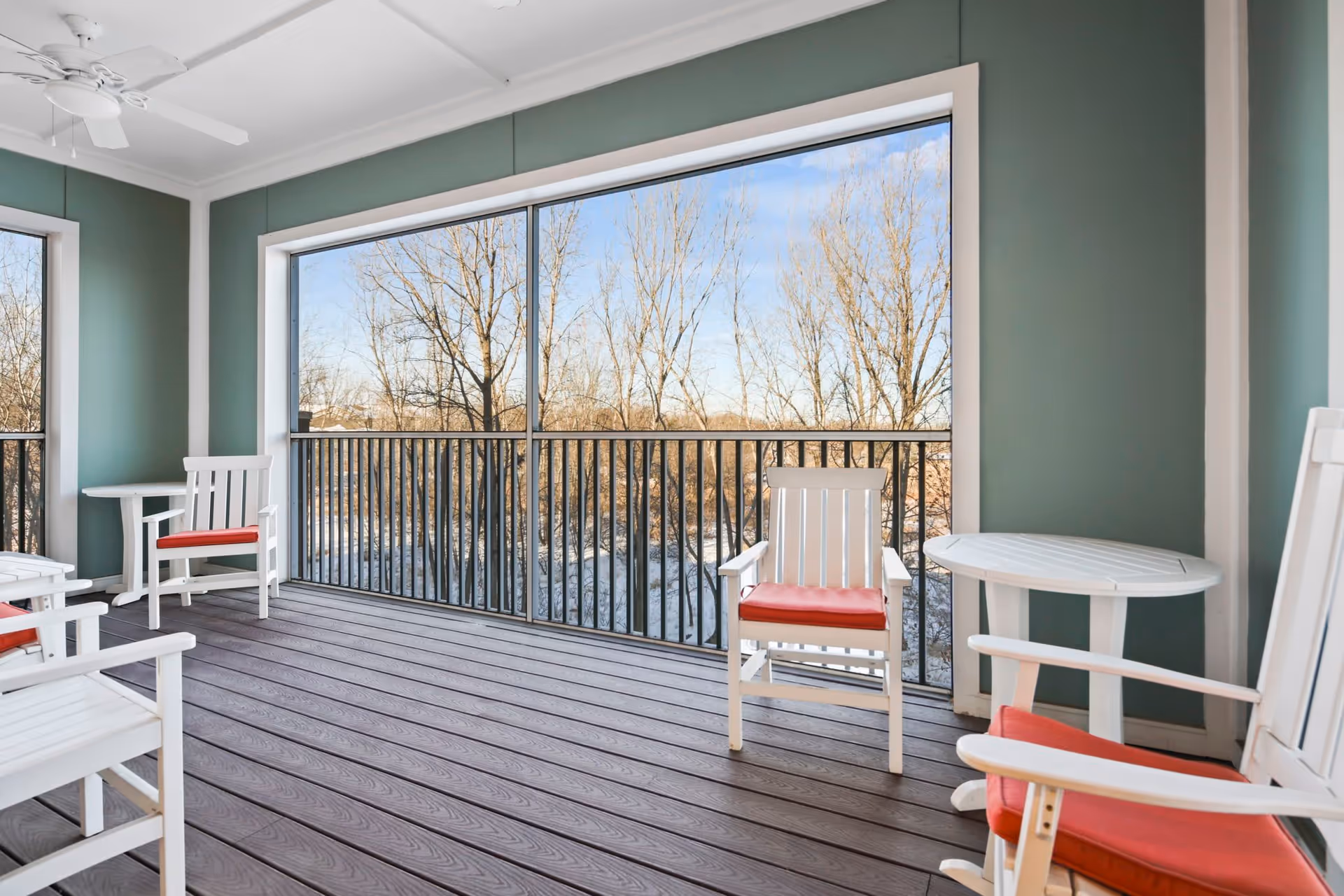 A screened-in porch with white wooden chairs featuring red cushions and small white tables. The porch has green walls, a ceiling fan, and overlooks leafless trees outside.