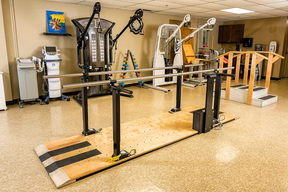 Interior therapy/exercise room with parallel walking bars, exercise machines, free weights and a small wooden step bridge.