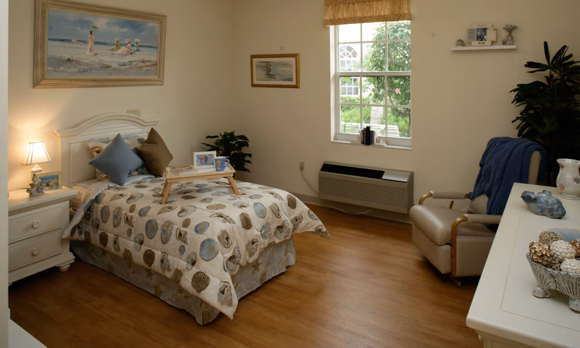 A cozy bedroom with a single bed covered in a seashell-patterned comforter and two pillows. A small wooden tray with a photo frame and a mug sits on the bed. To the left of the bed is a white nightstand with a lamp and decorative items. A large window with a beige valance lets in natural light, and a beige armchair with a blue throw blanket is positioned near a white dresser with decorative bowls and vases. The room has wooden flooring and light-colored walls adorned with framed artwork.