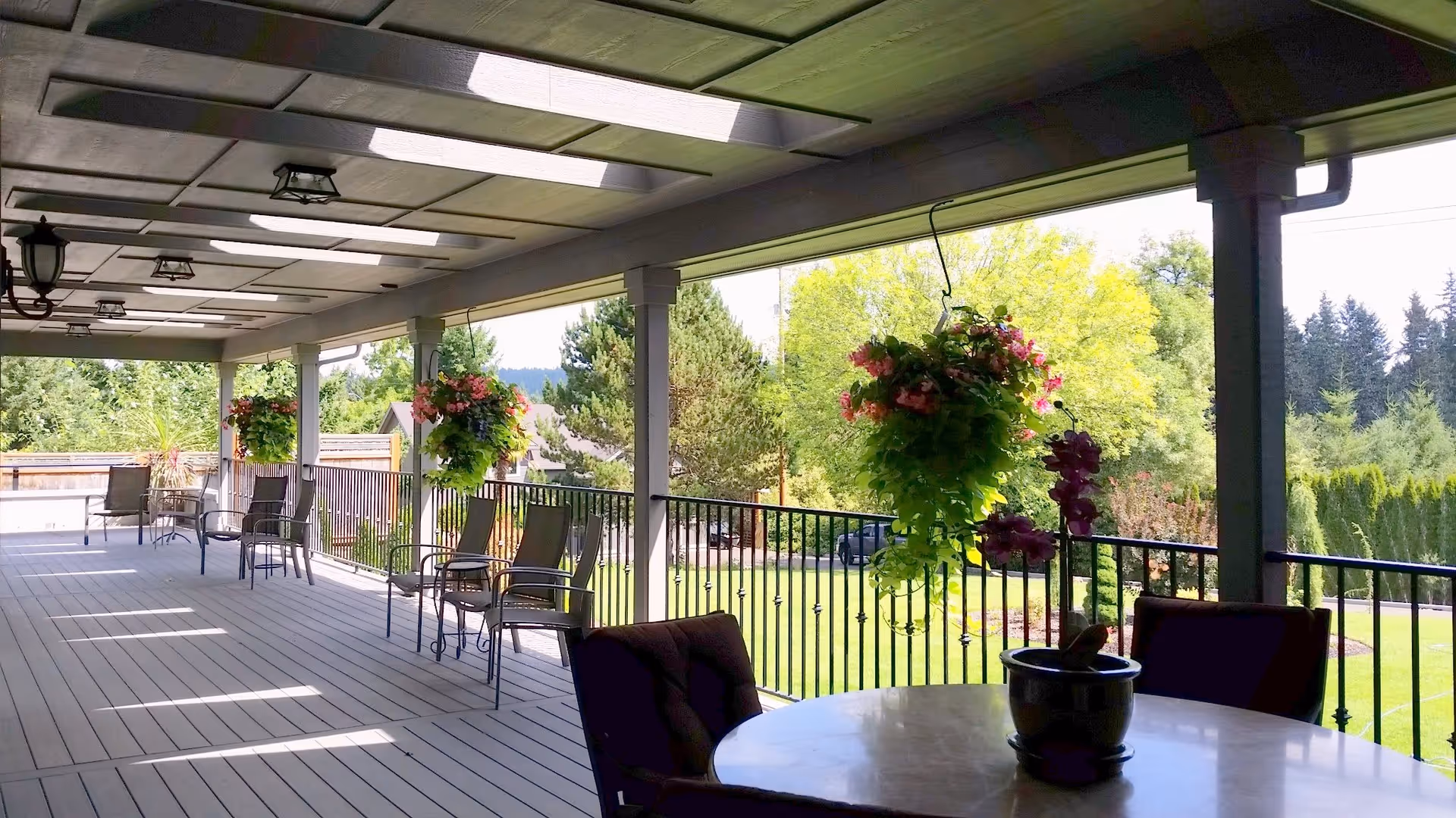 Covered outdoor patio area with several chairs and tables, hanging flower baskets, and a view of a green lawn and trees in the background.