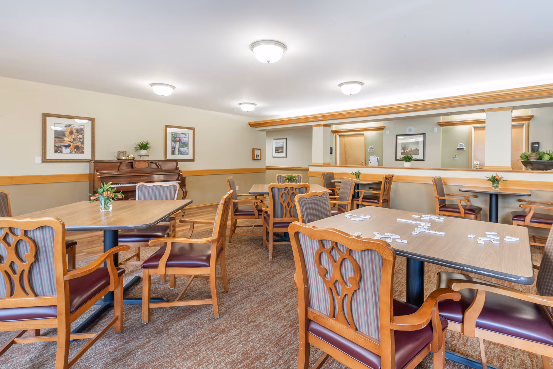 A well-lit common room with several wooden tables and chairs arranged for group activities. Some tables have small flower vases, and one table has game tiles spread out. There is a piano against the wall with framed pictures above it, and a large mirror on the right side reflecting part of the room.