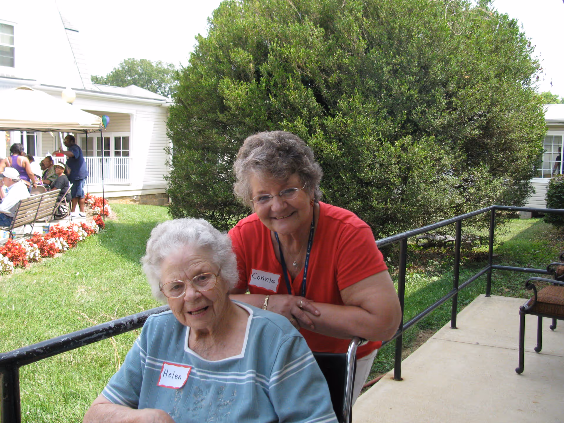 An elderly woman named Helen sitting in a wheelchair outside on a paved path next to a green lawn and bushes. Another woman named Connie is standing behind her, smiling and leaning on the wheelchair. In the background, there are other people sitting on benches and a white building with windows.