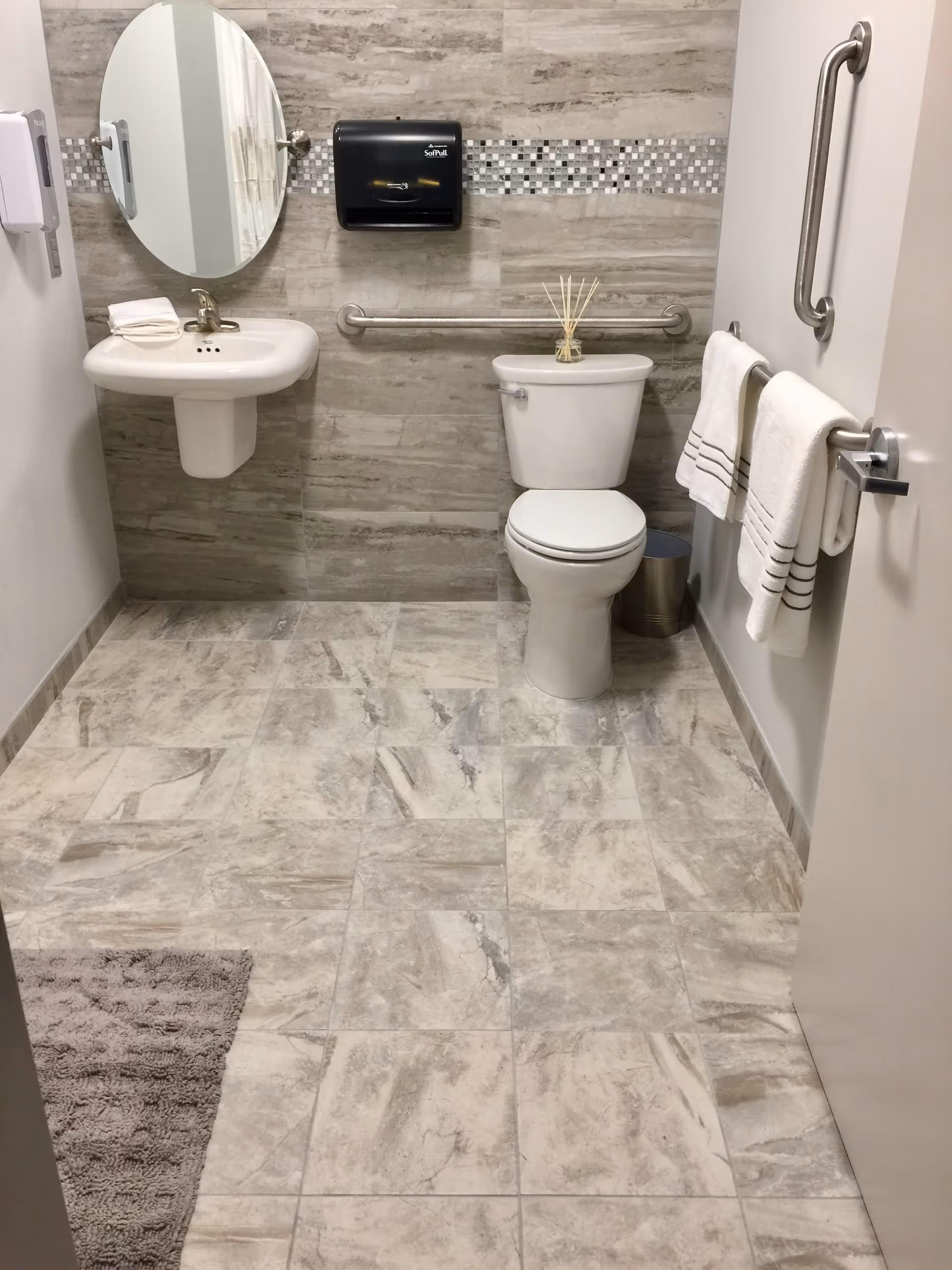 A clean and modern bathroom with beige and gray marble-patterned tile flooring and walls. The bathroom features a white toilet with a reed diffuser on top, a white wall-mounted sink with a folded towel, an oval mirror above the sink, a black paper towel dispenser, a stainless steel trash can, and two white towels hanging on a grab bar attached to the door. There are also grab bars on the wall near the toilet for accessibility.