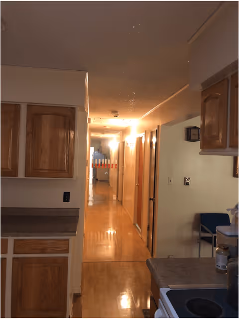 View of a hallway in a senior living facility with wooden cabinets and countertops on the left and right sides, a stove in the foreground on the right, and a chair against the wall in the background. The hallway is lit with ceiling lights and has wooden flooring.