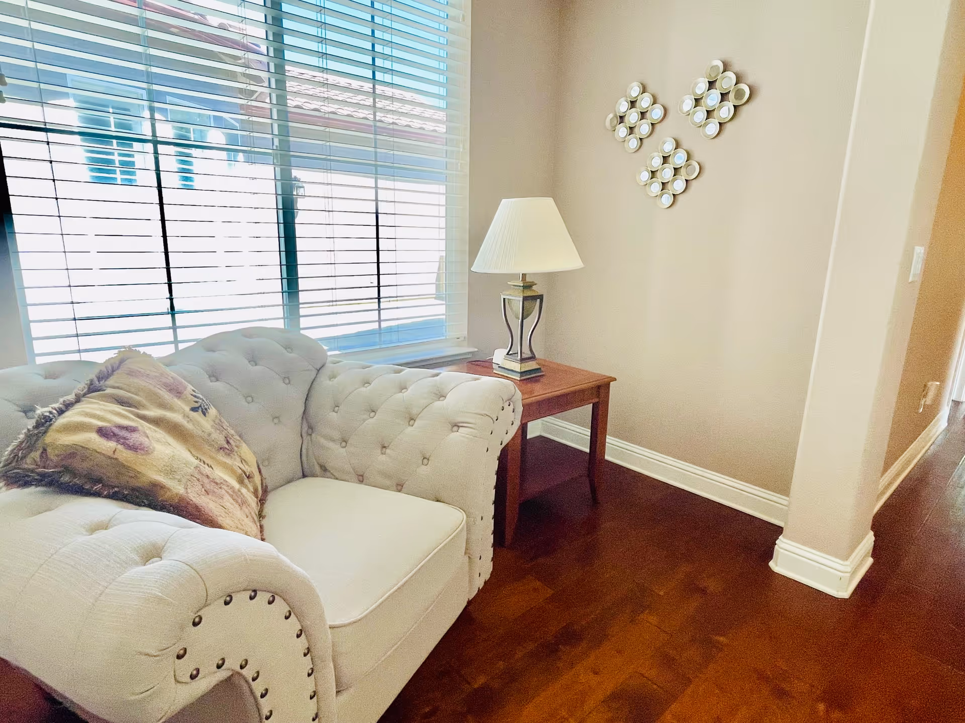 Bright living room corner with a tufted armchair, decorative pillow, wooden side table and lamp next to a window with blinds.