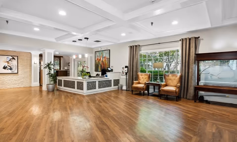 Spacious and well-lit reception area with wooden flooring, a white reception desk with decorative panels, two brown leather armchairs next to a window with curtains, a side table with a lamp, a large fish tank, and artwork on the walls.
