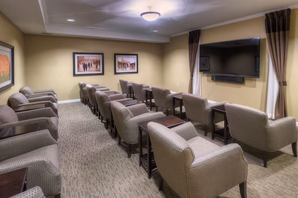 A small theater-style room with two rows of comfortable upholstered armchairs facing a large flat-screen TV mounted on the wall. The room has beige walls, carpeted floor, framed artwork on the walls, and windows with brown curtains on either side of the TV.