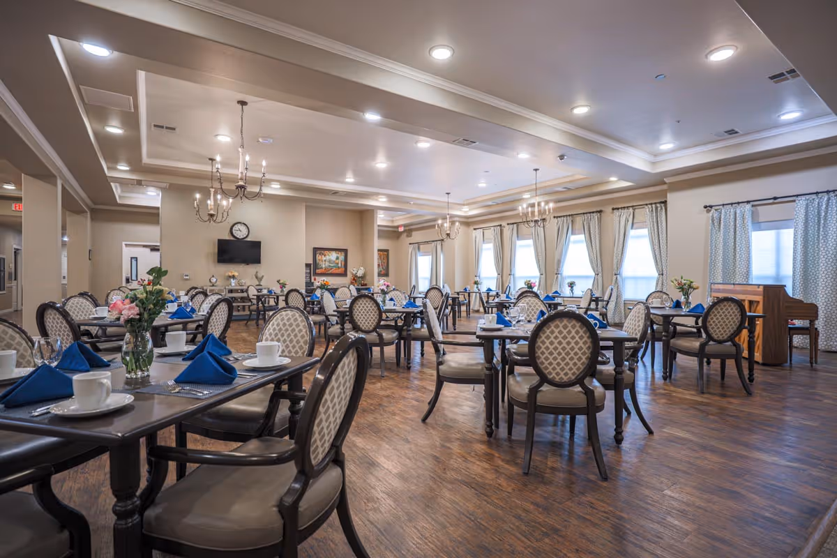 A spacious dining room in an assisted living facility with multiple tables set with white cups, blue napkins, and floral centerpieces. The room features elegant chairs with patterned upholstery, wooden flooring, chandeliers, large windows with curtains, and a piano in the corner.
