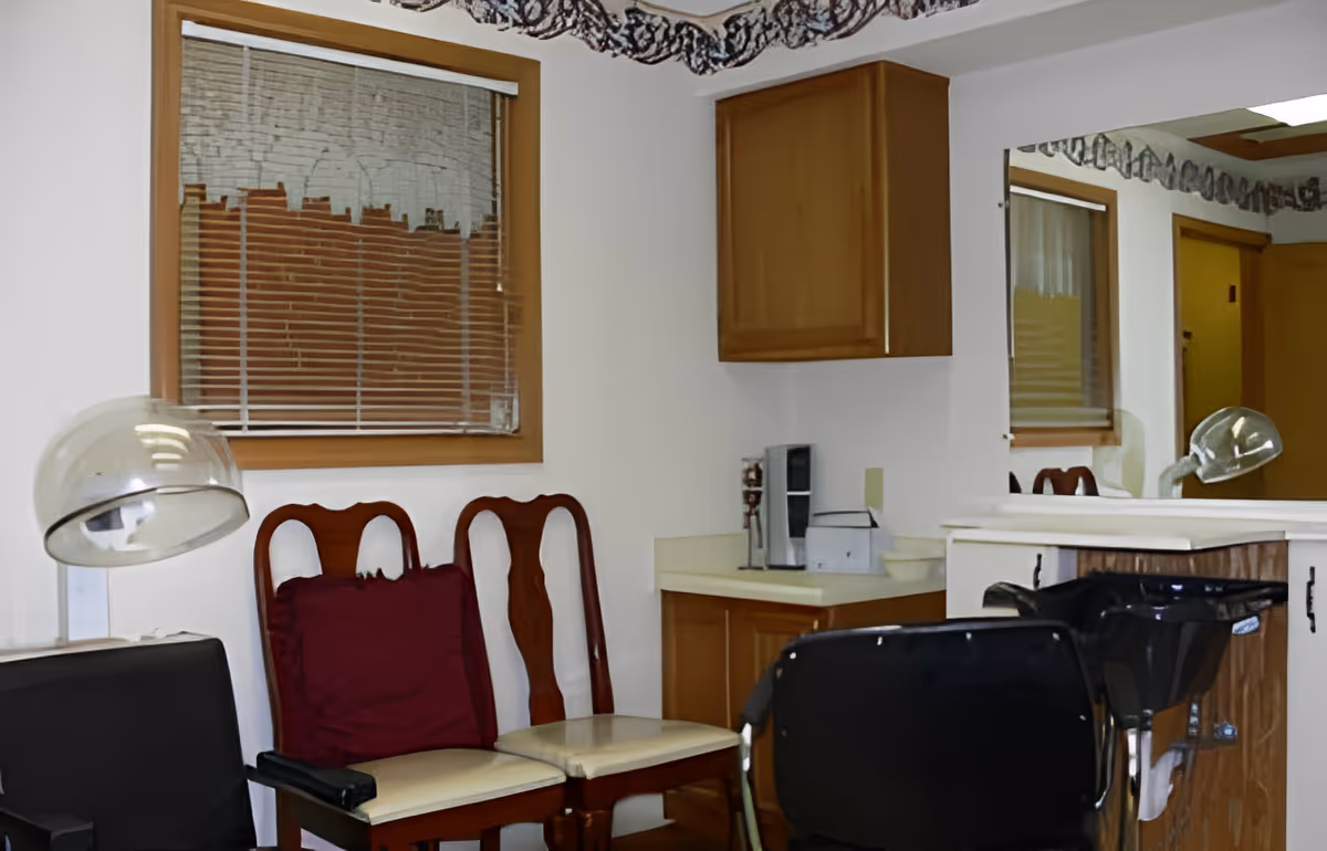 Interior of a small salon area with two wooden chairs with cream cushions and a maroon pillow, a black salon chair facing a black hair washing sink, a hair dryer with a clear hood, a window with closed blinds, wooden cabinets, a countertop with a coffee maker, and a large mirror reflecting part of the room.