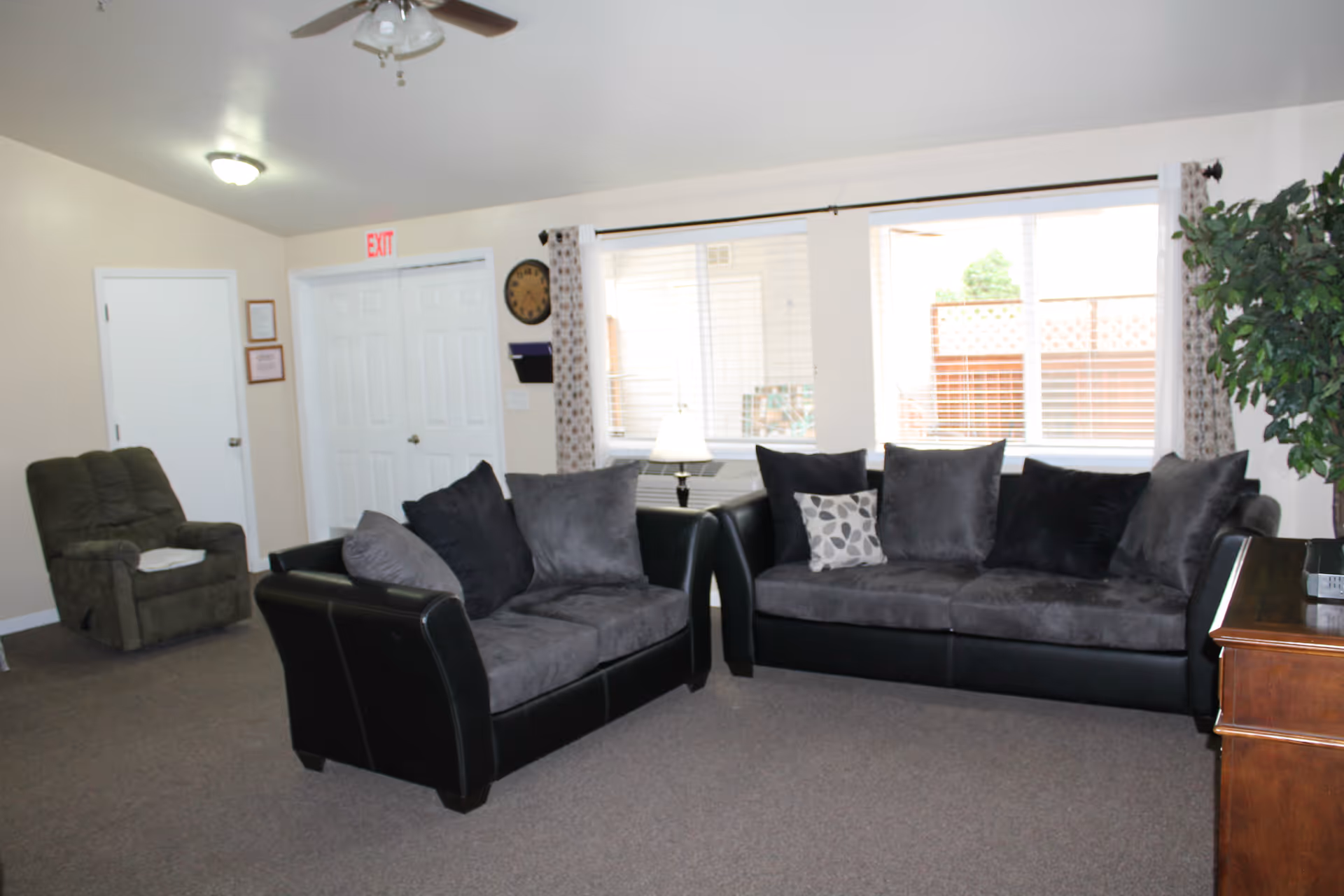 A living room with two black leather sofas with gray cushions, a green recliner chair, a wooden side table with a lamp, a large window with blinds and curtains, a ceiling fan with lights, and a potted plant in the corner.