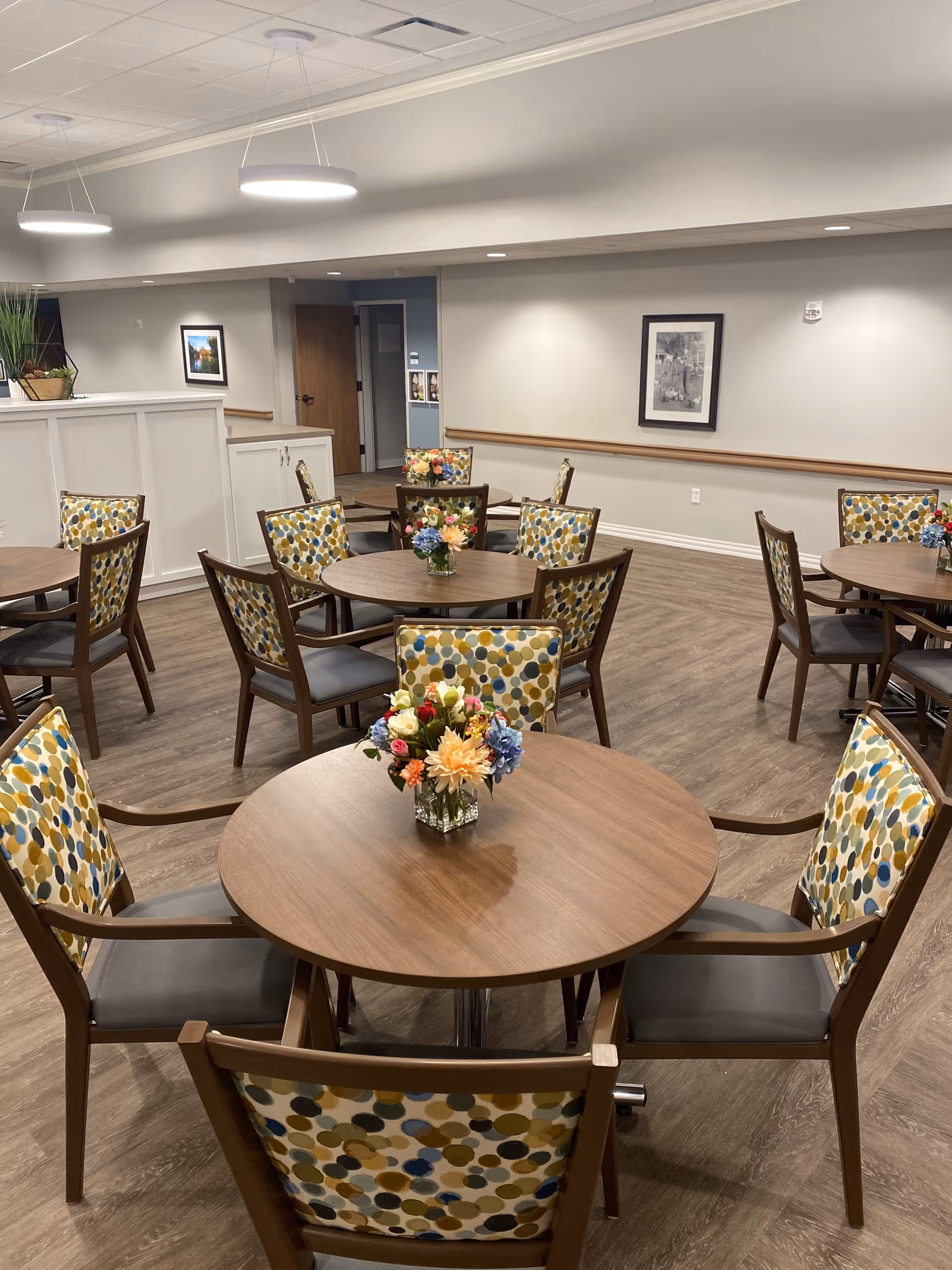 A well-lit dining area with round wooden tables and chairs featuring colorful polka dot upholstery. Each table has a small floral centerpiece. The room has light-colored walls, wood flooring, framed artwork on the walls, and modern ceiling lights.