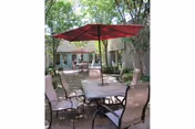 Outdoor patio area with a rectangular glass-top table surrounded by six cushioned chairs. A large red umbrella is centered over the table providing shade. The patio is surrounded by trees and greenery with a building visible in the background.