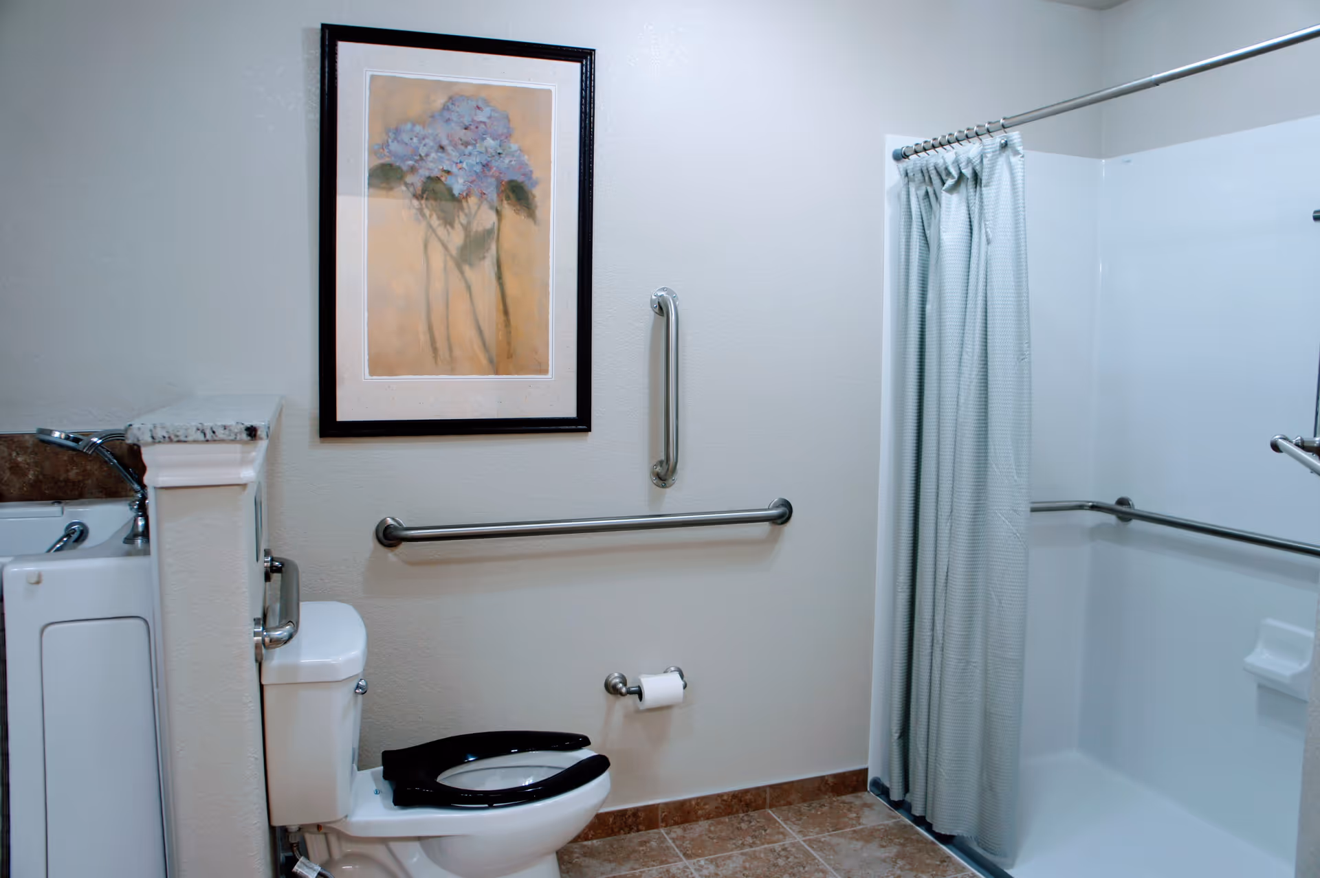 A senior living facility bathroom with a white toilet featuring a black seat, grab bars on the walls for support, a walk-in shower with a curtain, and a framed floral painting on the wall. The floor is tiled and the walls are light-colored.