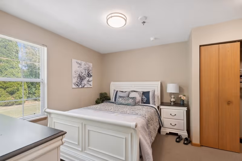 A neatly made bed with white wooden frame and multiple pillows in a bedroom with beige walls. There is a nightstand with a lamp and flowers next to the bed, a window with blinds showing greenery outside, a dresser, and a wooden closet door.