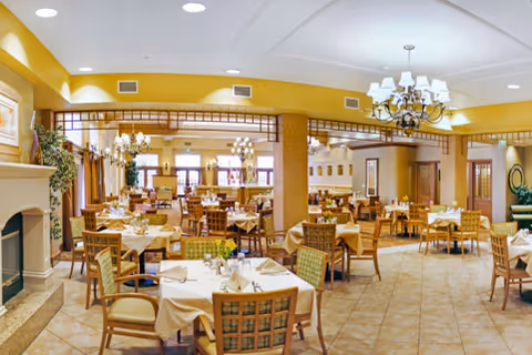 A spacious dining room with multiple tables covered with white tablecloths, set with glassware and napkins. The room features wooden chairs with patterned cushions, chandeliers hanging from the ceiling, large windows with curtains, and warm yellow walls. There is a fireplace on the left side and decorative plants near the windows.
