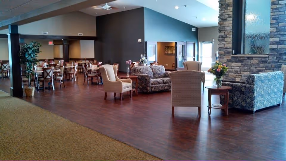 A spacious interior common area with wooden flooring and carpeted sections. The room features multiple seating arrangements including floral patterned sofas, beige armchairs, and wooden dining tables with chairs. There are flower vases on small tables and a stone accent wall with a water feature. The area is well-lit with natural light coming through windows and has a calm, welcoming atmosphere.
