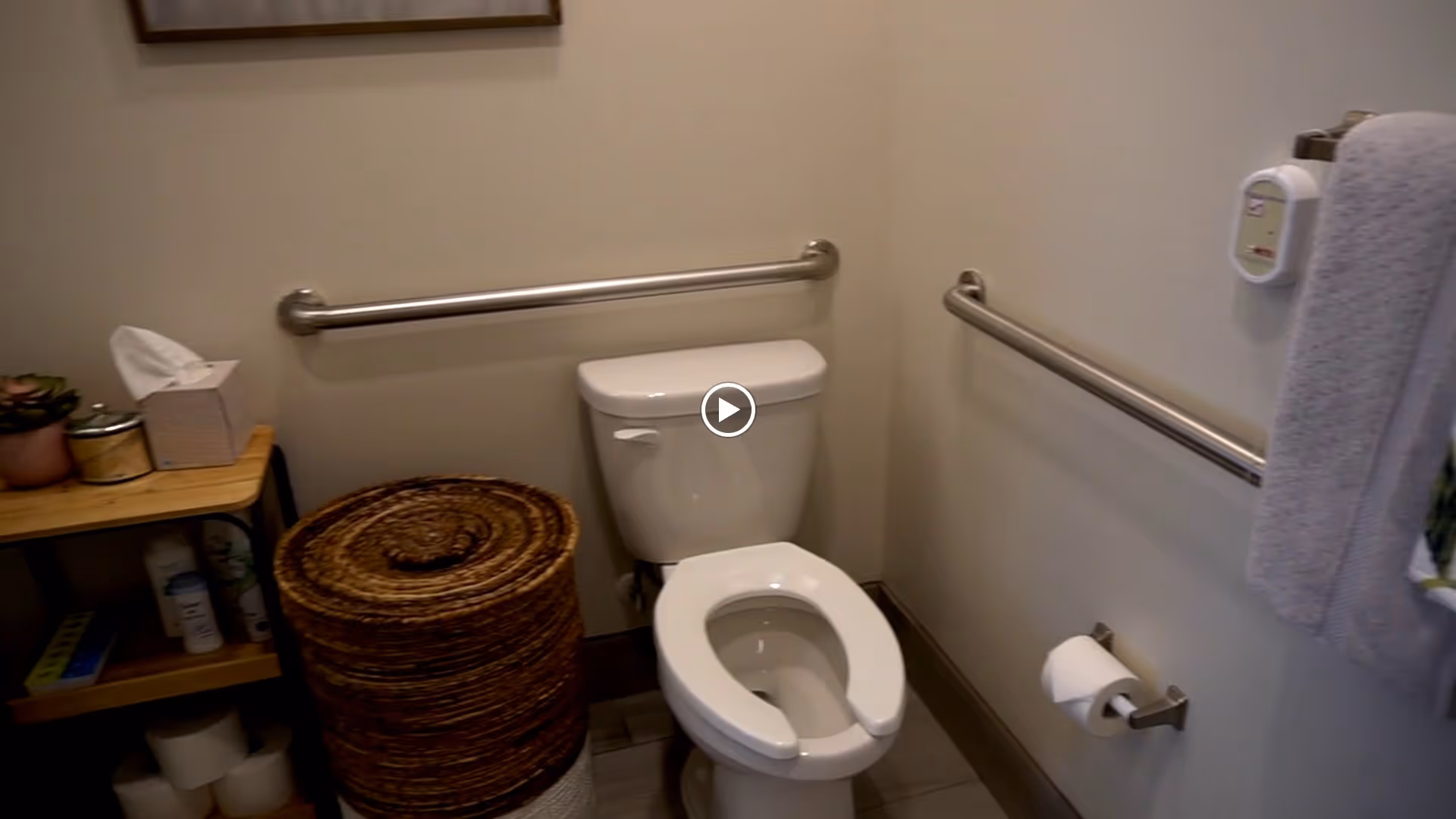 A small bathroom with a white toilet that has a raised toilet seat. There are metal grab bars on the walls behind and beside the toilet for support. To the left, there is a wooden shelf holding a tissue box, a small plant, and some toiletries. A woven basket is placed on the floor next to the shelf. On the right wall, a towel is hanging on a grab bar and a toilet paper holder is mounted below it.