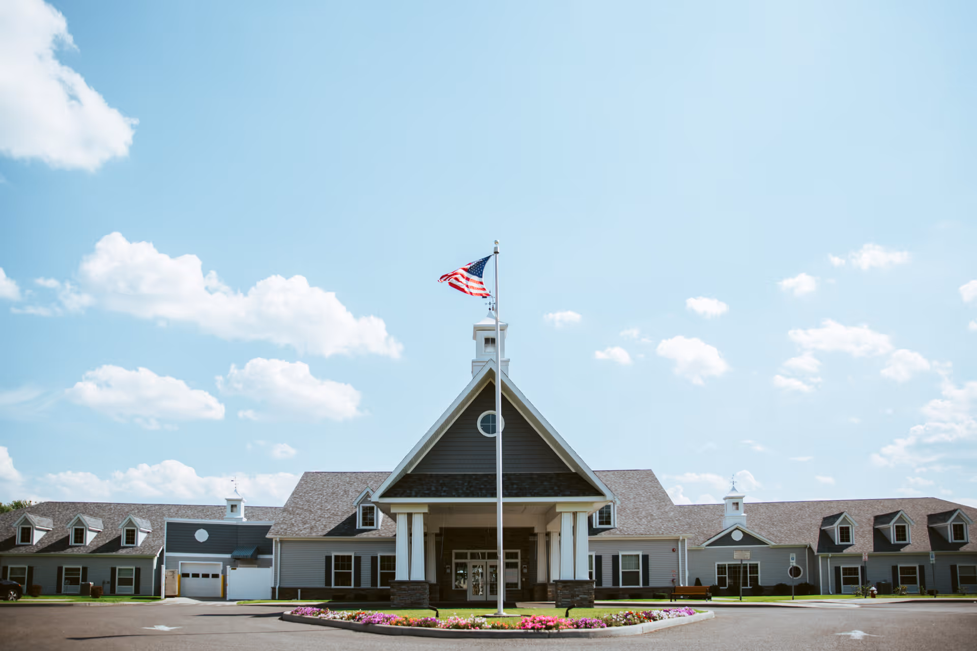 Front entrance of a senior living building with a flagpole flying an American flag and a circular flower bed.
