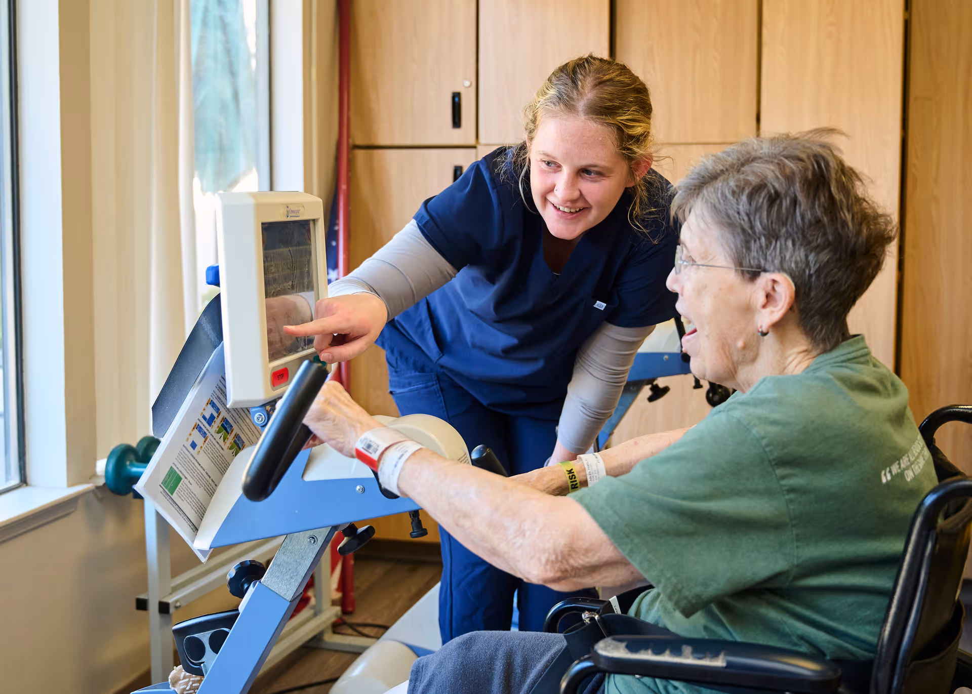 A healthcare worker in navy scrubs is assisting an elderly woman in a wheelchair with using a piece of exercise equipment that has a digital screen. They are indoors near a window with natural light coming in, and the healthcare worker is smiling and pointing at the screen while the elderly woman looks engaged.