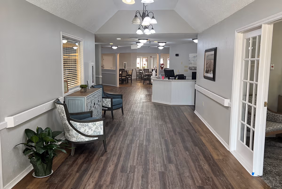 Interior hallway of Broadmoor Court facility with wood flooring, two upholstered chairs and a small cabinet on the left side, a potted plant on the floor, and a reception desk further down the hall. The hallway leads to a dining area with tables and chairs. The walls are painted light gray and there are multiple ceiling lights.