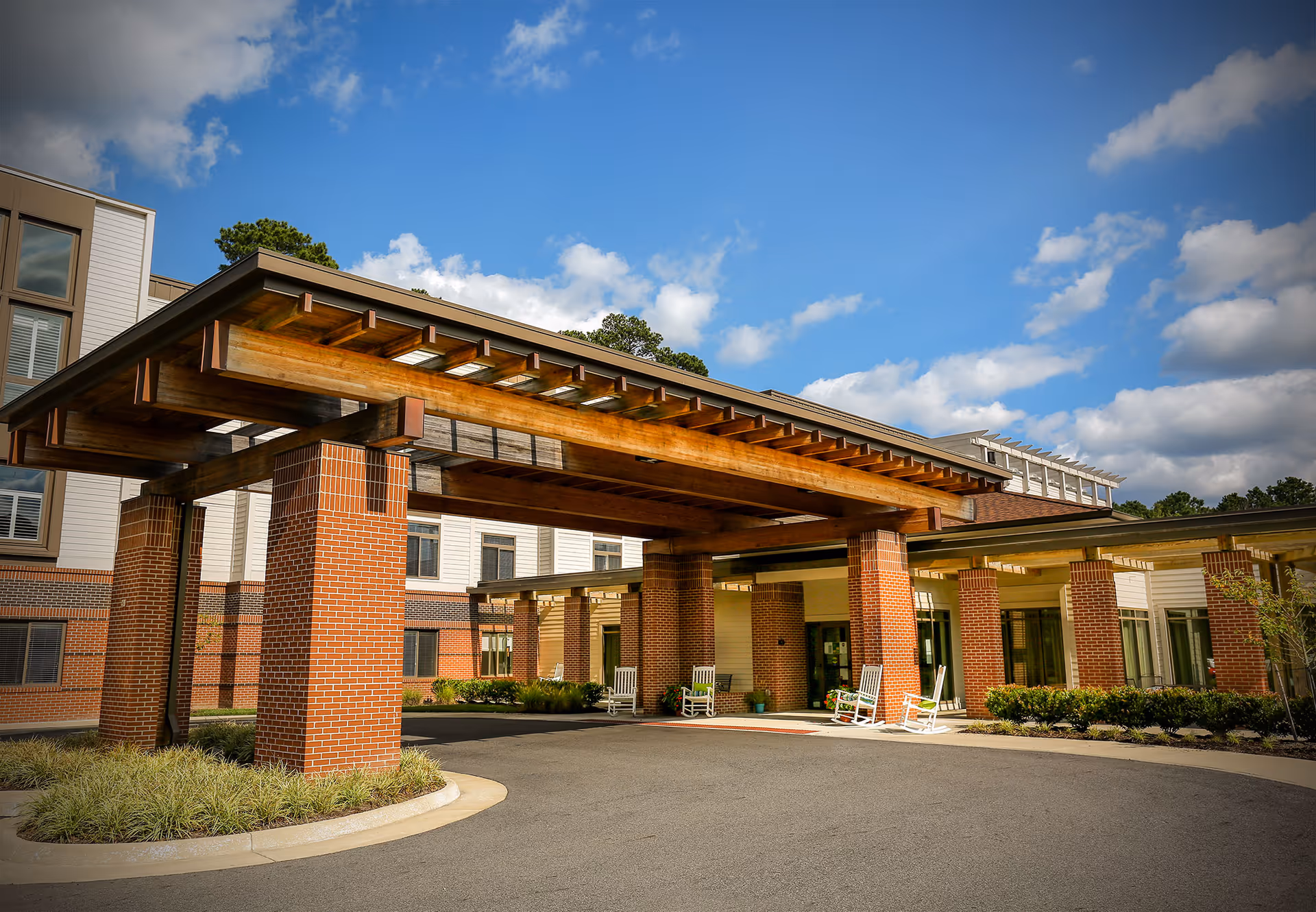Exterior view of The Hamilton Assisted Living facility showing a covered entrance with large wooden beams supported by brick pillars, a driveway, and several white rocking chairs near the entrance under a partly cloudy blue sky.