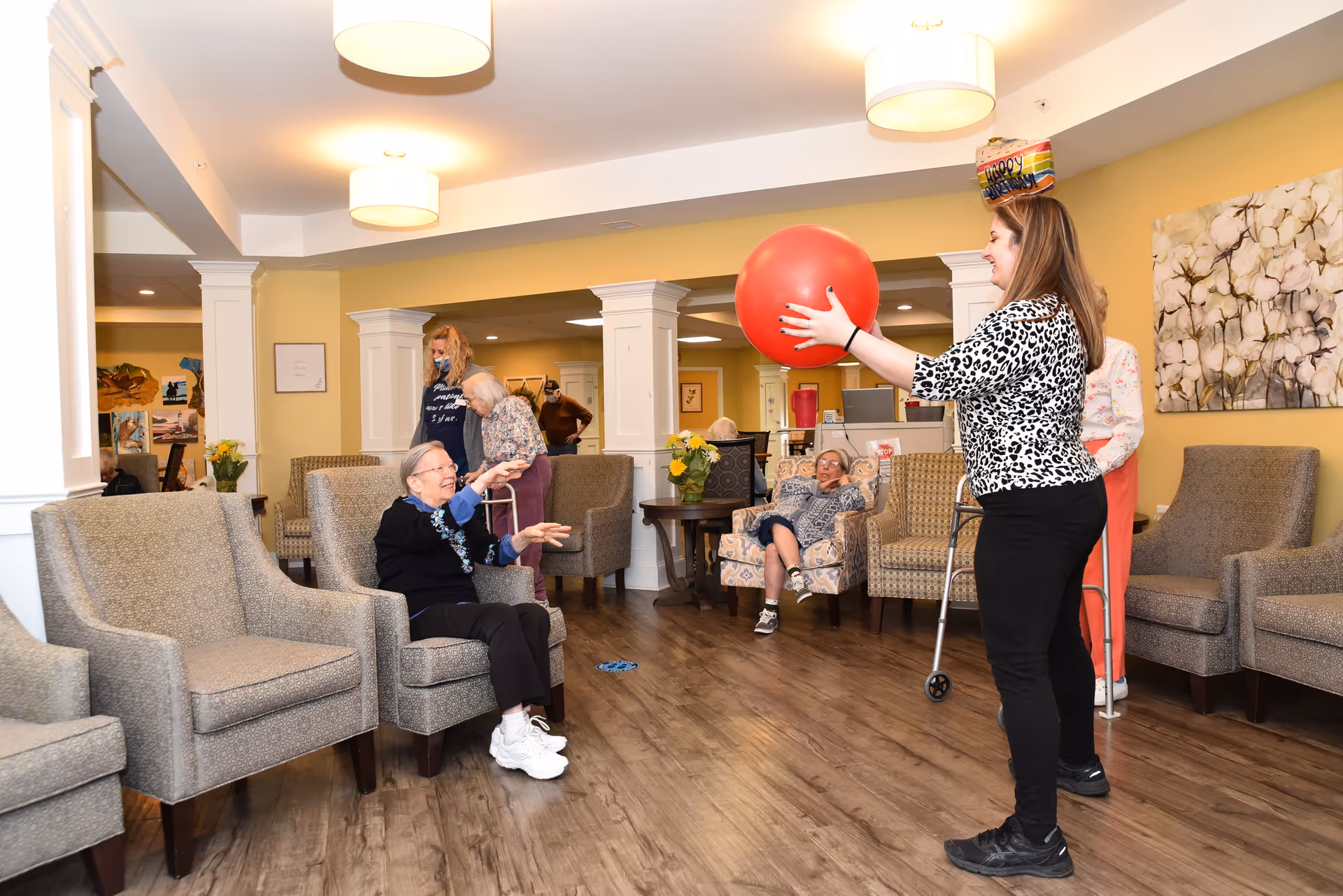 A group of elderly residents and a staff member are engaged in a recreational activity in a senior living facility common area. The staff member is holding a large red ball, preparing to toss it to a seated elderly woman who is reaching out to catch it. Other residents are seated in comfortable armchairs, watching and participating. The room has warm yellow walls, wooden flooring, and is decorated with flowers and artwork.