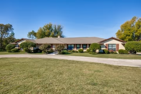 Single-story building with a beige exterior and brown roof surrounded by green bushes and trees under a clear blue sky, with a large grassy area in the foreground.