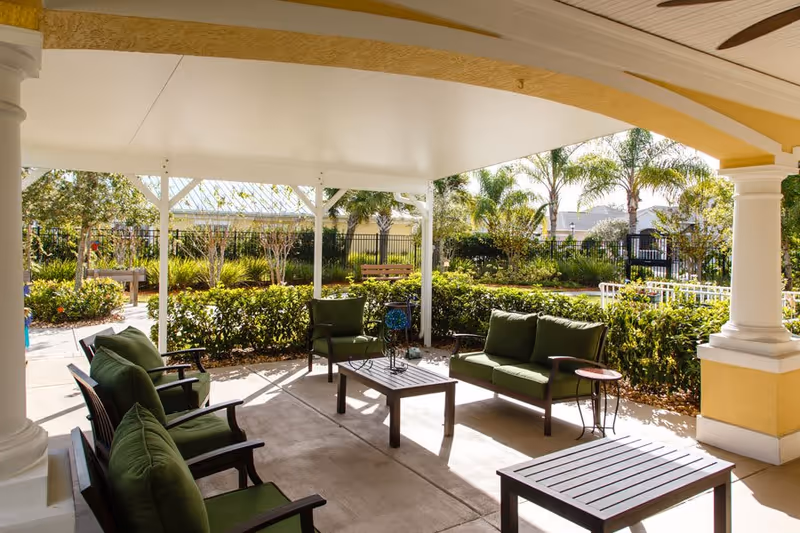 Covered outdoor patio with green-cushioned chairs and tables overlooking landscaped gardens.