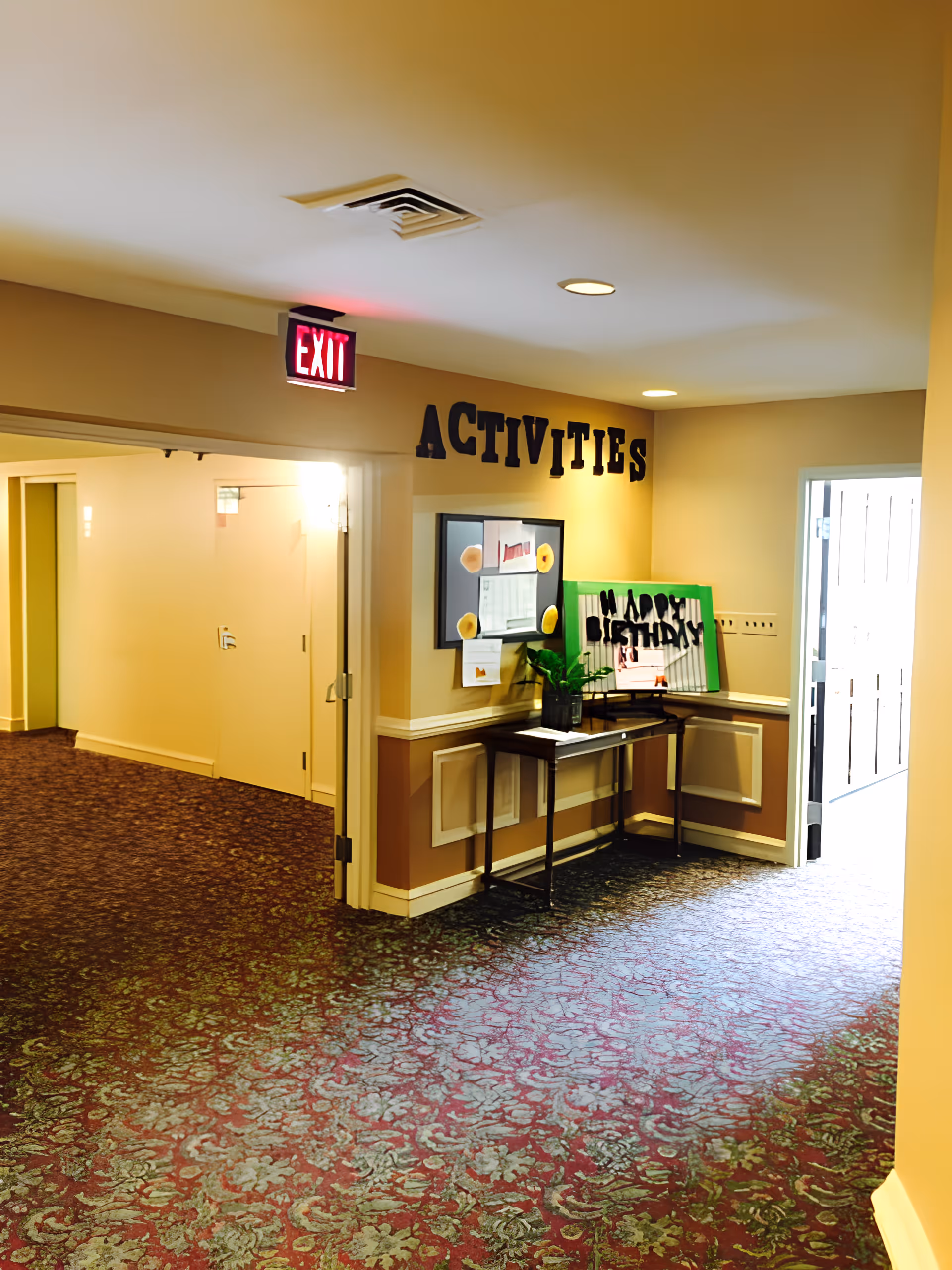 Interior hallway of Franklin Terrace Independent and Assisted Living with a patterned carpet and beige walls. There is a table against the wall with a plant and a sign that reads 'HAPPY BIRTHDAY'. Above the table, the word 'ACTIVITIES' is displayed on the wall. An exit sign is visible above a doorway leading to another area.