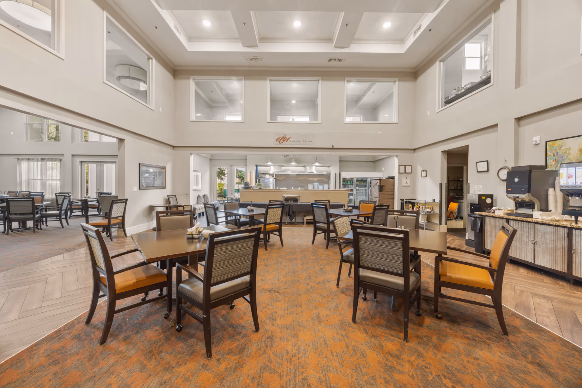 A spacious dining area in a senior living facility with multiple tables and chairs arranged on an orange patterned carpet. The room has high ceilings with large windows near the ceiling, allowing natural light to enter. In the background, there is a kitchen area with stainless steel appliances and a counter. To the right, there is a beverage station with a coffee machine and cups.