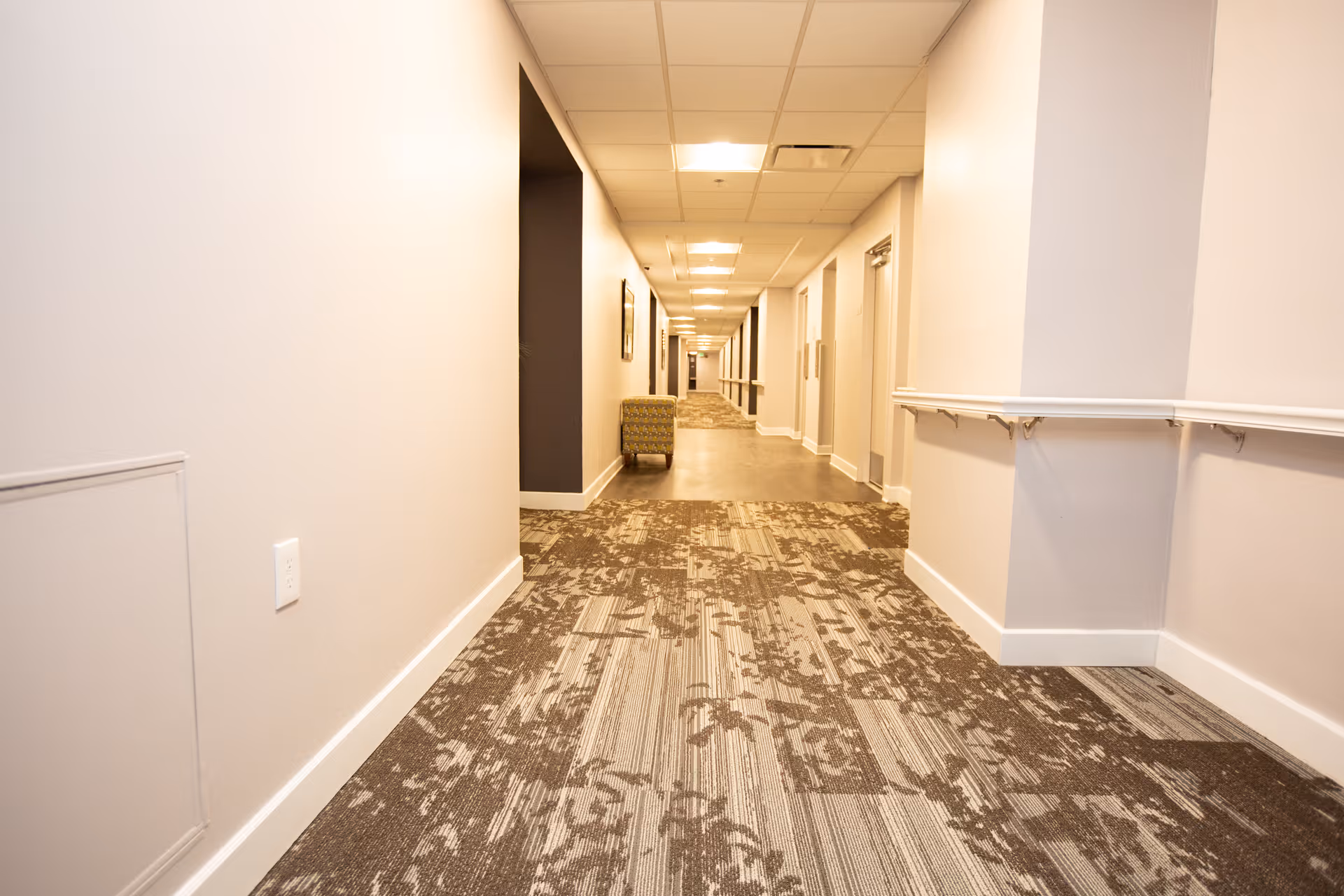 A long, well-lit hallway in a senior living facility with patterned carpet flooring, white walls, ceiling lights, and a handrail on the right side. There is a single armchair placed along the hallway and several doors on the right wall.