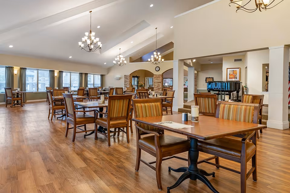 Spacious dining room with wooden floors, multiple tables and chairs arranged neatly, large windows with curtains allowing natural light, chandeliers hanging from a vaulted ceiling, and a black grand piano in the background.