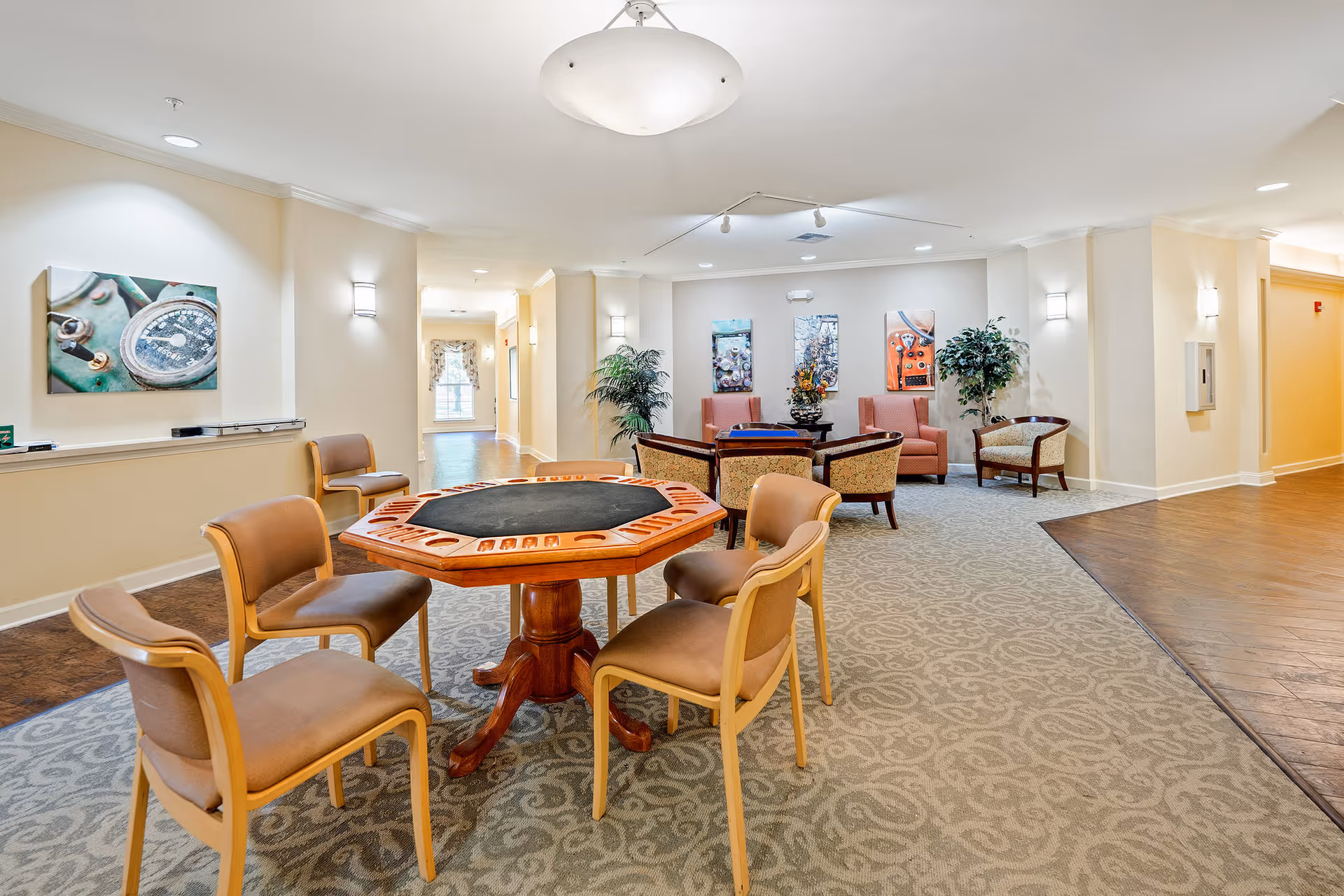 A well-lit common area in a senior living facility featuring a wooden octagonal game table surrounded by four cushioned chairs. In the background, there are additional seating arrangements with armchairs and a small table with a floral centerpiece. The walls are decorated with colorful artwork and there are potted plants adding greenery to the space. The floor is carpeted with a patterned design, and the ceiling has recessed lighting and a central light fixture.