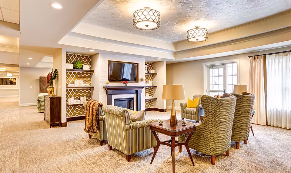 A bright and spacious living room area in a senior living facility with patterned armchairs and a sofa arranged around a wooden coffee table and a fireplace with a mounted flat-screen TV above it. The room features built-in shelves with decorative items, large windows with sheer curtains, and ceiling light fixtures.